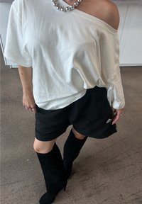 Balta per didelė marškiniai su off-shoulder dizainu, derinami su juodomis šortais, ant kurių matomas Adidas logotipas. Juodi iki kelių ilgio batai užbaigia įvaizdį.
