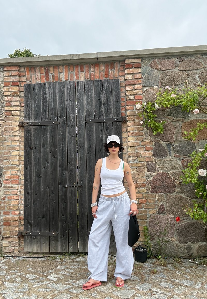 Personne portant un débardeur blanc, un pantalon large gris, une casquette blanche, des lunettes de soleil et des sandales roses se tient devant une grande porte en bois avec des murs en briques et en pierre.