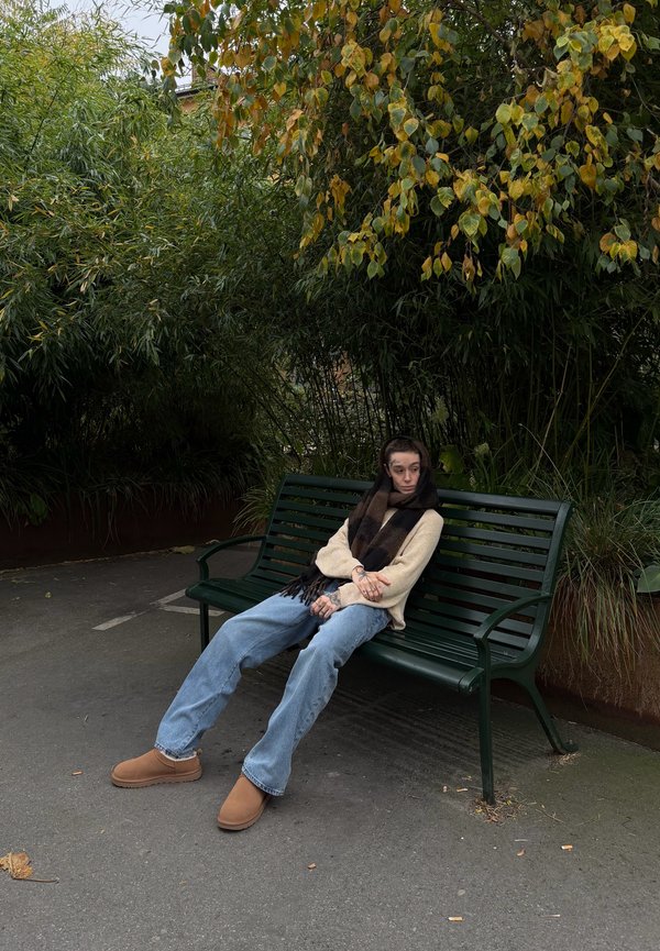Persona con bufanda, suéter beige, vaqueros y zapatos marrones sentada en un banco verde del parque bajo árboles frondosos.