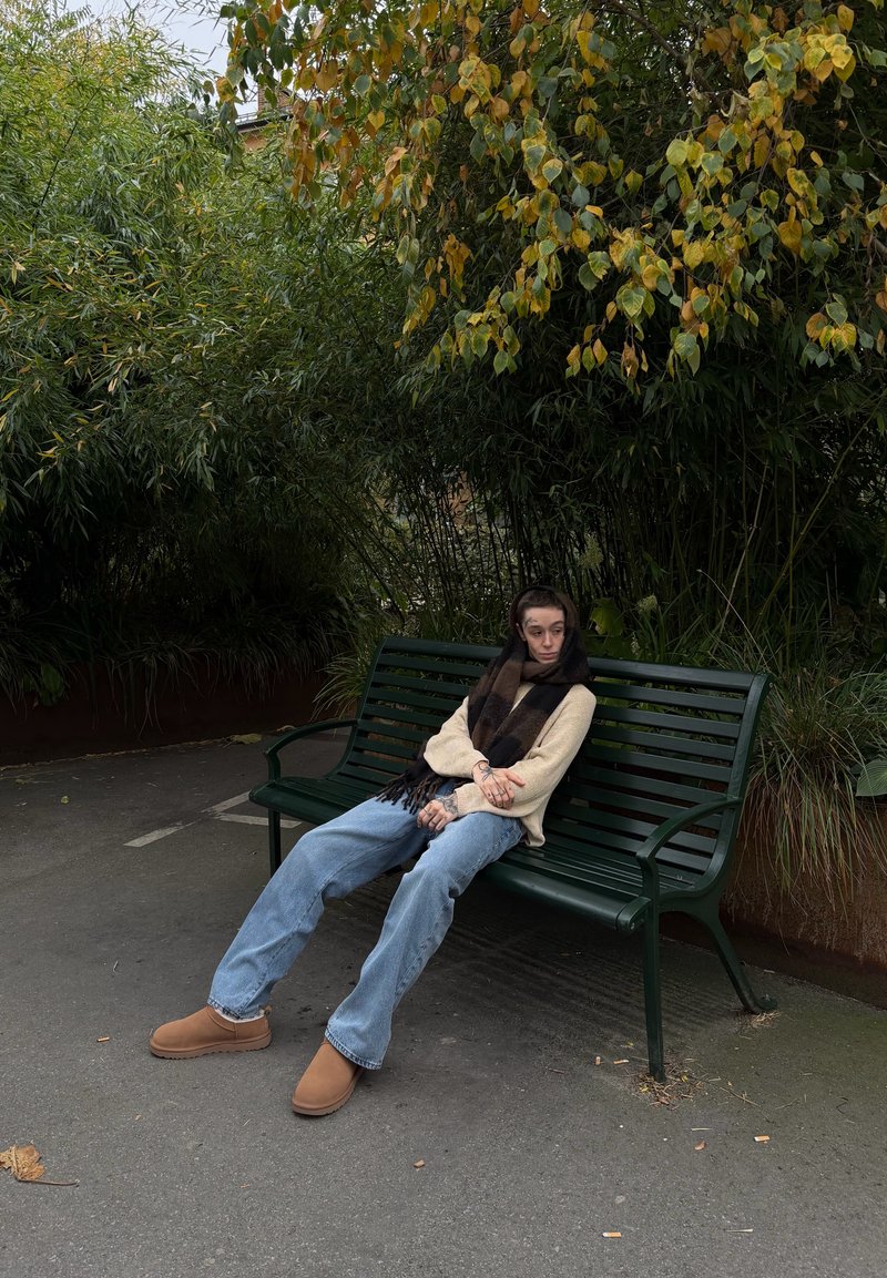Persona con bufanda, suéter beige, vaqueros y zapatos marrones sentada en un banco verde del parque bajo árboles frondosos.