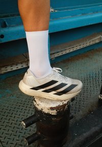 Kāja baltā zeķē ar baltu Adidas kedu ar melnbaltām svītrām, balstīta uz nolietota metāla staba, uz industriāla grīdas fona.