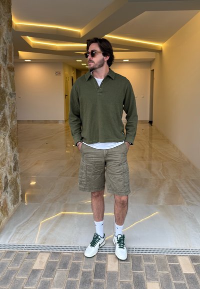 Polo de manga larga verde oliva, pantalones cortos cargo caqui, zapatillas blancas con detalles en verde, de pie en un interior moderno y bien iluminado.