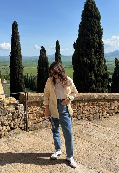 Mujer vestida con una camiseta a rayas amarilla clara sobre una camiseta blanca, jeans azules claros, sosteniendo un bolso transparente, de pie sobre un pavimento de piedra.