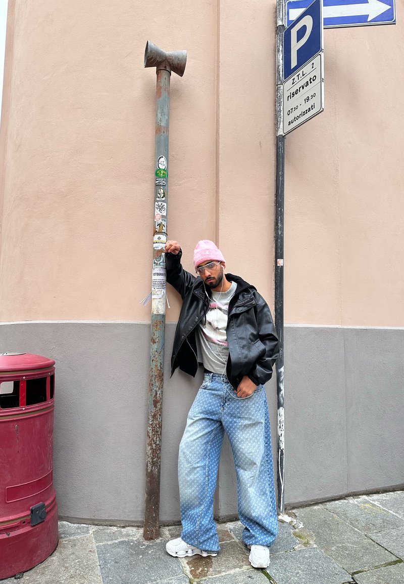 Homme portant un bonnet rose et une veste en cuir noire, appuyé sur un poteau rouillé couvert d'autocollants, debout à un coin de rue urbain près d'un panneau de stationnement et d'une poubelle rouge.