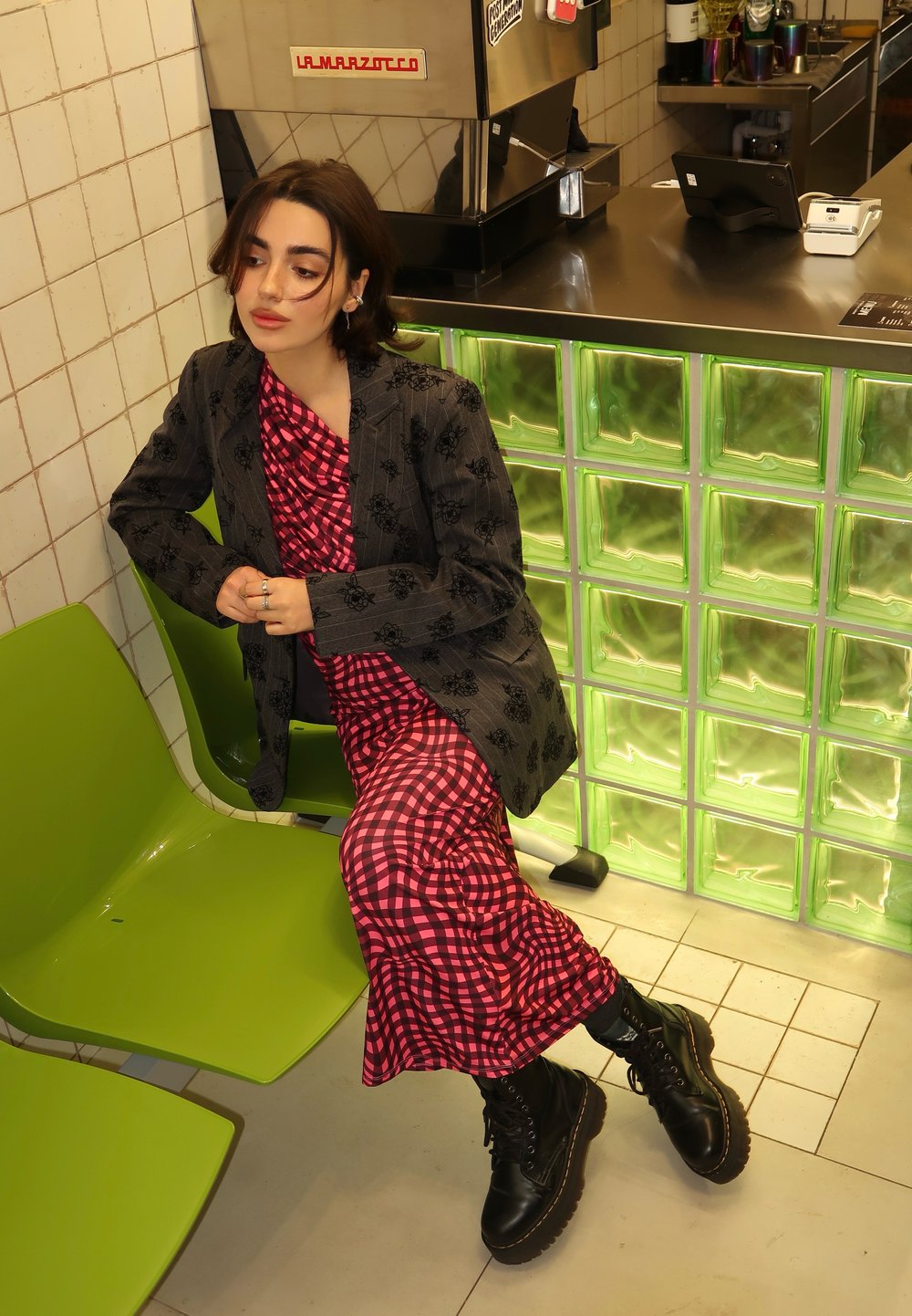 Zwart patroon blazer over een roze geruite jurk, zwarte combat laarzen, zittend op een groene stoel tegen een glazen blokmuur.