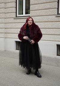 Άτομο με κόκκινα μαλλιά που φοράει μπορντό γούνινο μπουφάν, μακριά μαύρη φούστα από τούλι και μαύρα παπούτσια με πλατφόρμα, στέκεται σε πεζοδρόμιο της πόλης.