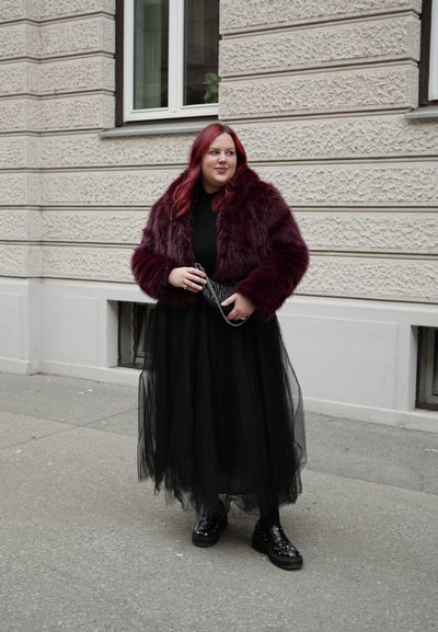 Persona con cabello rojo que lleva una chaqueta de piel sintética burdeos, una falda larga de tul negro y zapatos de plataforma negros, de pie en una acera de la ciudad.
