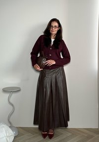 Βουργουνδί κάρντιγκαν, λευκή μπλούζα, καφέ πλισέ δερμάτινη φούστα, καφέ τσάντα χειρός με λεπτομέρειες από μεταλλικό υλικό, και κόκκινα ψηλοτάκουνα με尖尖отов στην ανοιχτόχρωμη ξύλινη επιφάνεια.