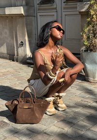 Bolso tote de ante marrón con un cierre dorado, pantalones cortos blancos, top beige y zapatillas multicolor. Los accesorios incluyen pulseras apiladas y gafas de sol.