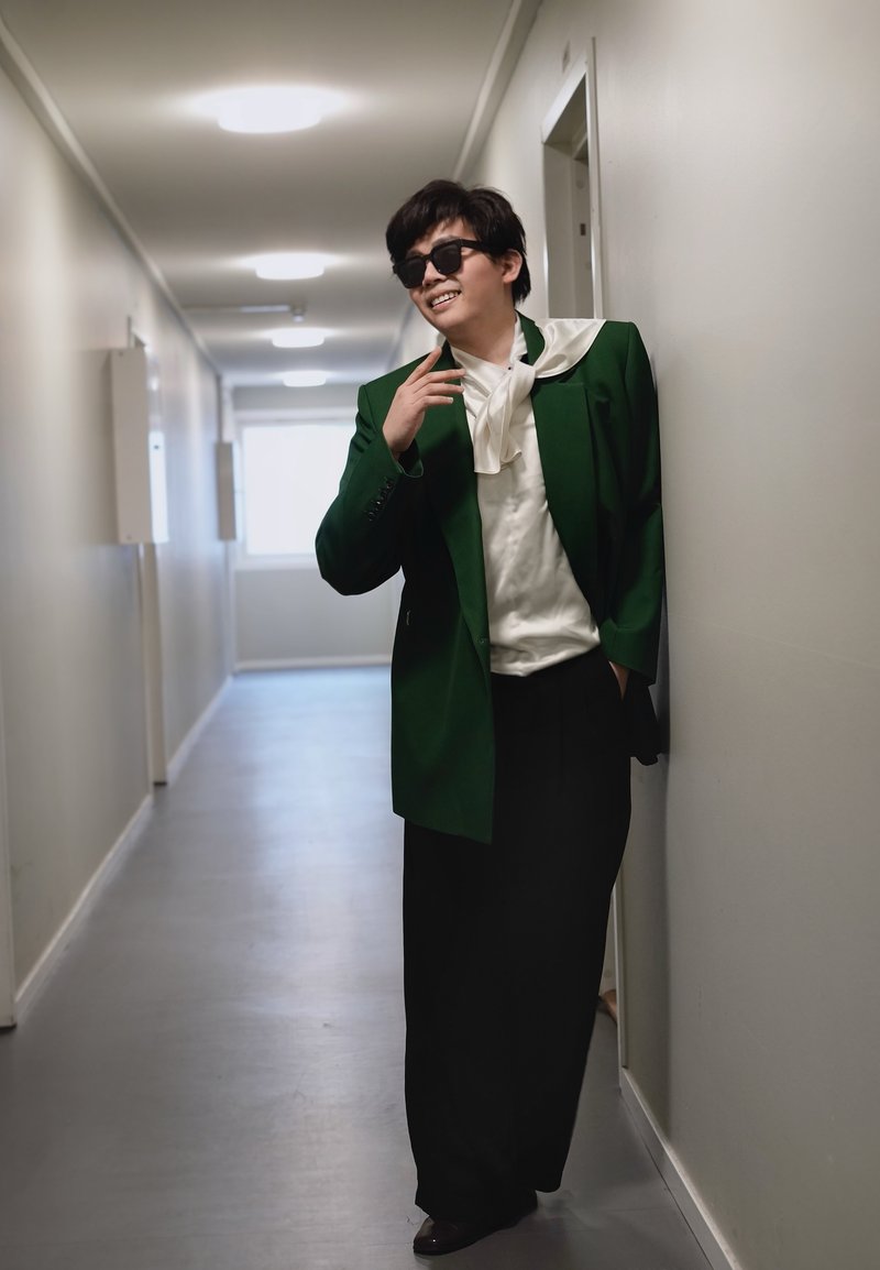 Homme portant des lunettes de soleil foncées, un blazer vert et un chemisier blanc, appuyé contre un mur de corridor, souriant avec une main levée près du visage.