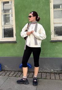 Balta mezgimo cardigan su dryžuota šaliku, derinama su juodomis trumpomis kelnių ir tamsiais sportbačiais. Fone matoma žalia siena ir langas.