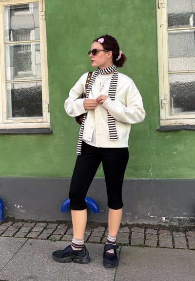 Cárdigan blanco de punto sobre una bufanda a rayas, combinado con pantalones negros recortados y zapatillas oscuras. El fondo presenta una pared verde y una ventana.