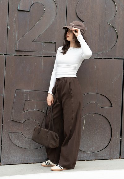 Pantalones marrones de pierna ancha y una camiseta blanca de manga larga ajustada, combinados con una gorra marrón y un bolso marrón, complementan unas zapatillas claras con rayas.