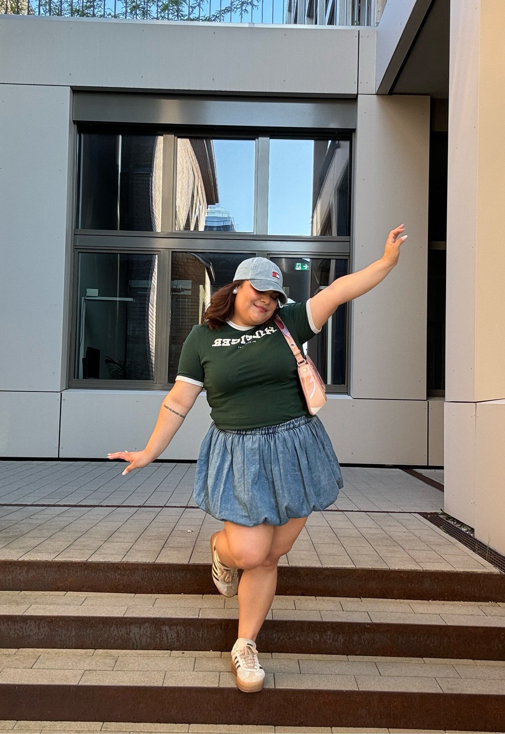 Πράσινο κοντομάνικο πουκάμισο με λευκές λεπτομέρειες, τζιν φούστα, γκρίζο καπέλο και μπεζ πλατφόρμες, ποζάροντας σε σκαλοπάτια σε μια σύγχρονη αστική περιοχή.