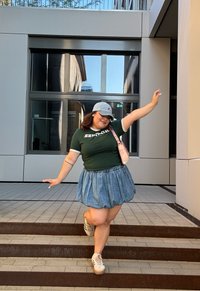 Πράσινο κοντομάνικο πουκάμισο με λευκές λεπτομέρειες, τζιν φούστα, γκρίζο καπέλο και μπεζ πλατφόρμες, ποζάροντας σε σκαλοπάτια σε μια σύγχρονη αστική περιοχή.