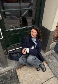 Temno moder volnen plašč čez svetel pulover, sive hlače in črni škornji. Sedi na tleh ob zeleni vratih in črni torbi.