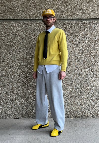 Suéter amarillo sobre una camisa blanca, corbata negra, pantalones de chándal grises y zapatillas amarillas y negras. Lleva una gorra amarilla con "LOCAL SPACE".