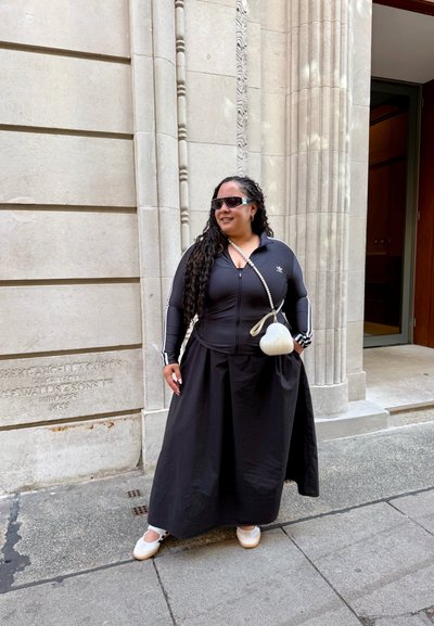 Chaqueta deportiva negra con cremallera y rayas blancas, combinada con una falda larga negra. Lleva un bolso blanco y unas gafas de sol de gran tamaño.