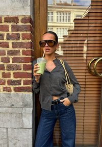 Cardigan grigio con bottoni, jeans blu a gamba larga, occhiali da sole marroni oversize, tiene in mano una bevanda verde, si trova contro un muro di mattoni con dettagli in legno.