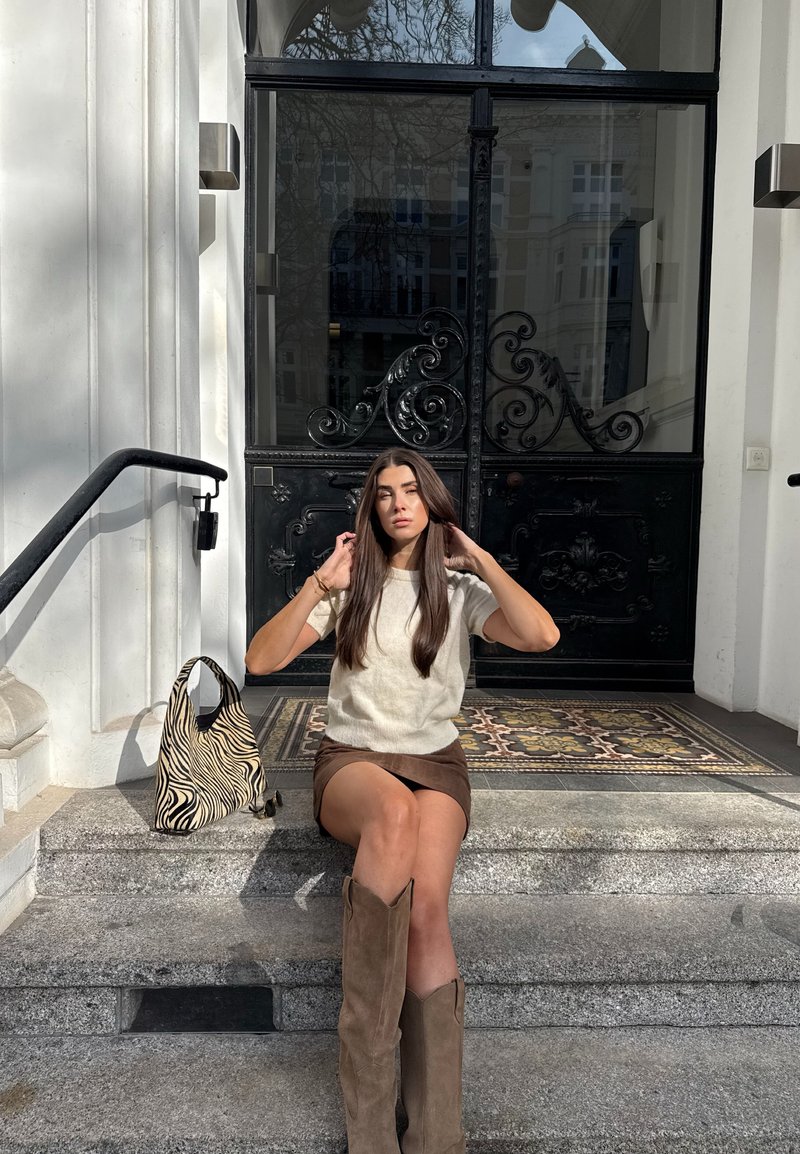 Νεαρή γυναίκα που ρυθμίζει τα μαλλιά της ενώ κάθεται σε πέτρινα σκαλιά έξω από μια μαύρη, περίτεχνη πόρτα, με μια τσάντα με σχέδιο ζέβρας δίπλα της.