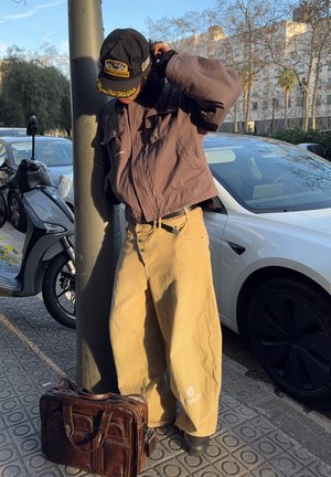 Persona con chaqueta marrón de gran tamaño y pantalones beige ajusta una gorra negra mientras se apoya en un poste de la calle; bolsa de cuero marrón en la acera cerca.