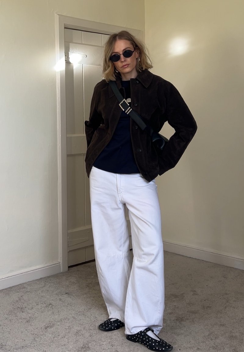 Frau trägt schwarze Jacke, marineblaues Top, weiße weite Hosen, schwarze Schuhe mit Punkten, schwarze Schultertasche und runde schwarze Sonnenbrille, steht drinnen.