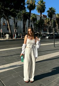 Cropped Top in lino bianco e pantaloni larghi, abbinati a una camicia bianca trasparente, mentre tiene una bottiglia d'acqua verde. Palme e edifici sullo sfondo.