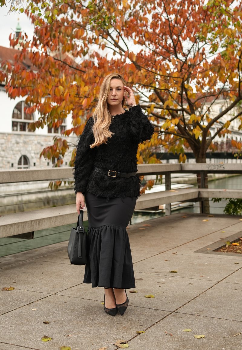 Schwarzer Pullover mit strukturierter Oberfläche, langärmlig und mit rundem Ausschnitt, kombiniert mit einem schwarzen ausgestellten Rock und spitzen schwarzen High Heels. Schwarze Handtasche.