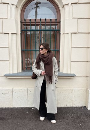 Gabardina gris claro sobre pantalones negros, con una bufanda marrón oscuro. Gafas de sol negras y zapatos blancos sin cordones. Edificio de ladrillo al fondo.