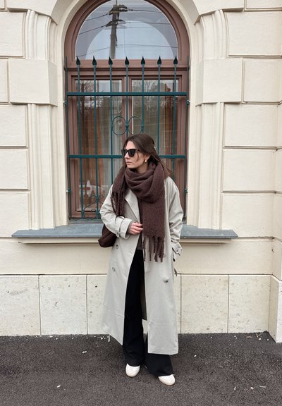 Gabardina gris claro sobre pantalones negros, con una bufanda marrón oscuro. Gafas de sol negras y zapatos blancos sin cordones. Edificio de ladrillo al fondo.