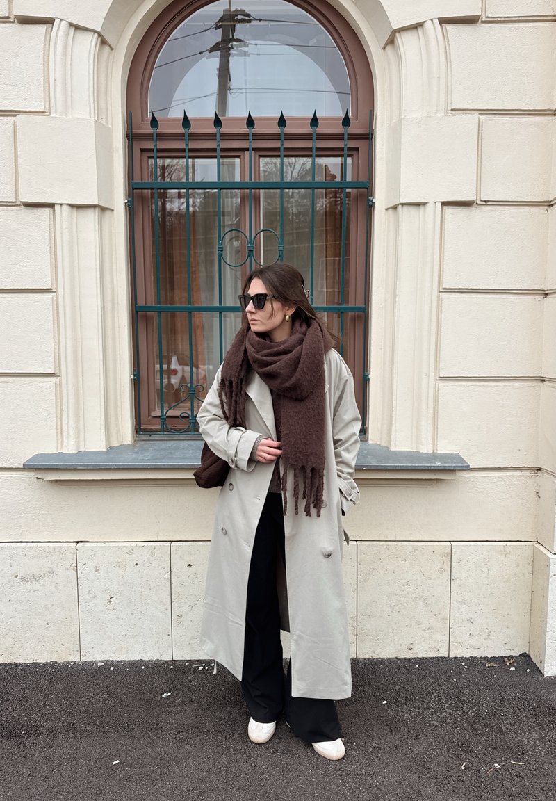 Hellgrauer Trenchcoat über schwarzen Hosen, mit einem dunkelbraunen Schal. Schwarze Sonnenbrille und weiße Slip-On-Schuhe. Im Hintergrund ein Backsteingebäude.
