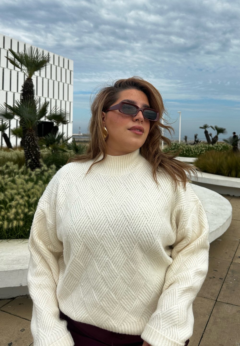 Χοντρή κρεμ ζακέτα με διαμαντέ πατρόν, ψηλό λαιμό και oversized μανίκια, συνδυασμένη με σκούρα ορθογώνια γυαλιά ηλίου.