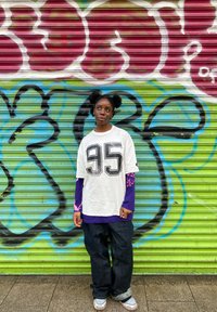 T-shirt branca oversize com estampagem preta "95", usada sobre uma camisola de manga comprida roxa. Jeans escuros e ténis cinzentos contra um fundo colorido de graffiti.