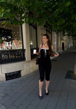 Camiseta negra ajustada con acentos blancos y cintas elásticas, combinada con leggings negros recortados. Tacones negros de charol y un bolso burdeos. Entorno en la acera.