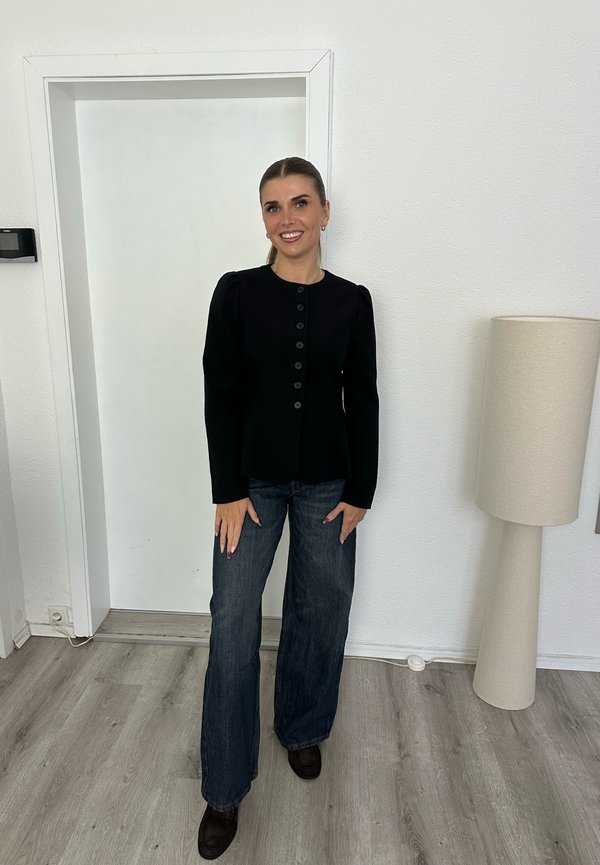 Femme debout à l'intérieur, portant une veste noire boutonnée, un jean large et des chaussures foncées, souriant avec les cheveux attachés vers l'arrière, à côté d'une grande lampe de sol beige.
