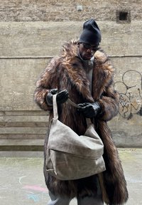Homme portant un manteau en fourrure, des gants noirs, un bonnet et des lunettes de soleil ajustant la sangle d'un grand sac en cuir beige à l'extérieur devant un mur de graffiti.