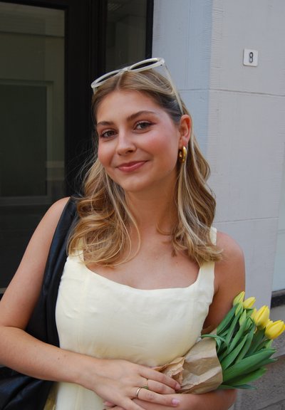 Vestido de tela amarillo claro con escote cuadrado, llevando un ramo de tulipanes amarillos envuelto en papel marrón. Bolso negro sobre el hombro.