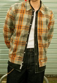 Chemise à carreaux aux tons verts, oranges et bleus, design à boutons avec des accents noirs, et tissu texturé. Présente un logo de marque visible.