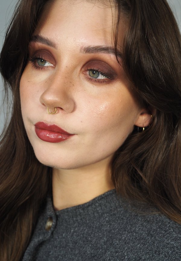 Look de maquillage avec ombre à paupières marron chaud, lèvres nude polies et piercing septum doré. Fond neutre, avec des ombres douces.