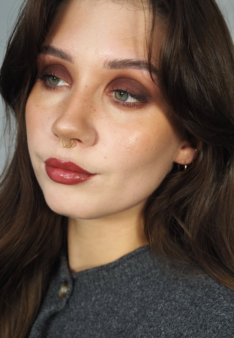 Trucco con ombretto marrone caldo, labbra nude lucide e piercing al setto dorato. Sfondo neutro, con ombre morbide.