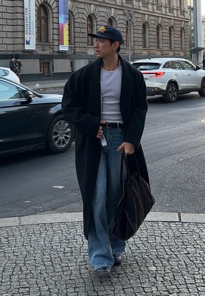 Abrigo negro de gran tamaño, camisa gris y jeans de corte amplio. Los accesorios incluyen un bolso marrón oscuro y una gorra azul marino con un logo dorado. Entorno urbano.