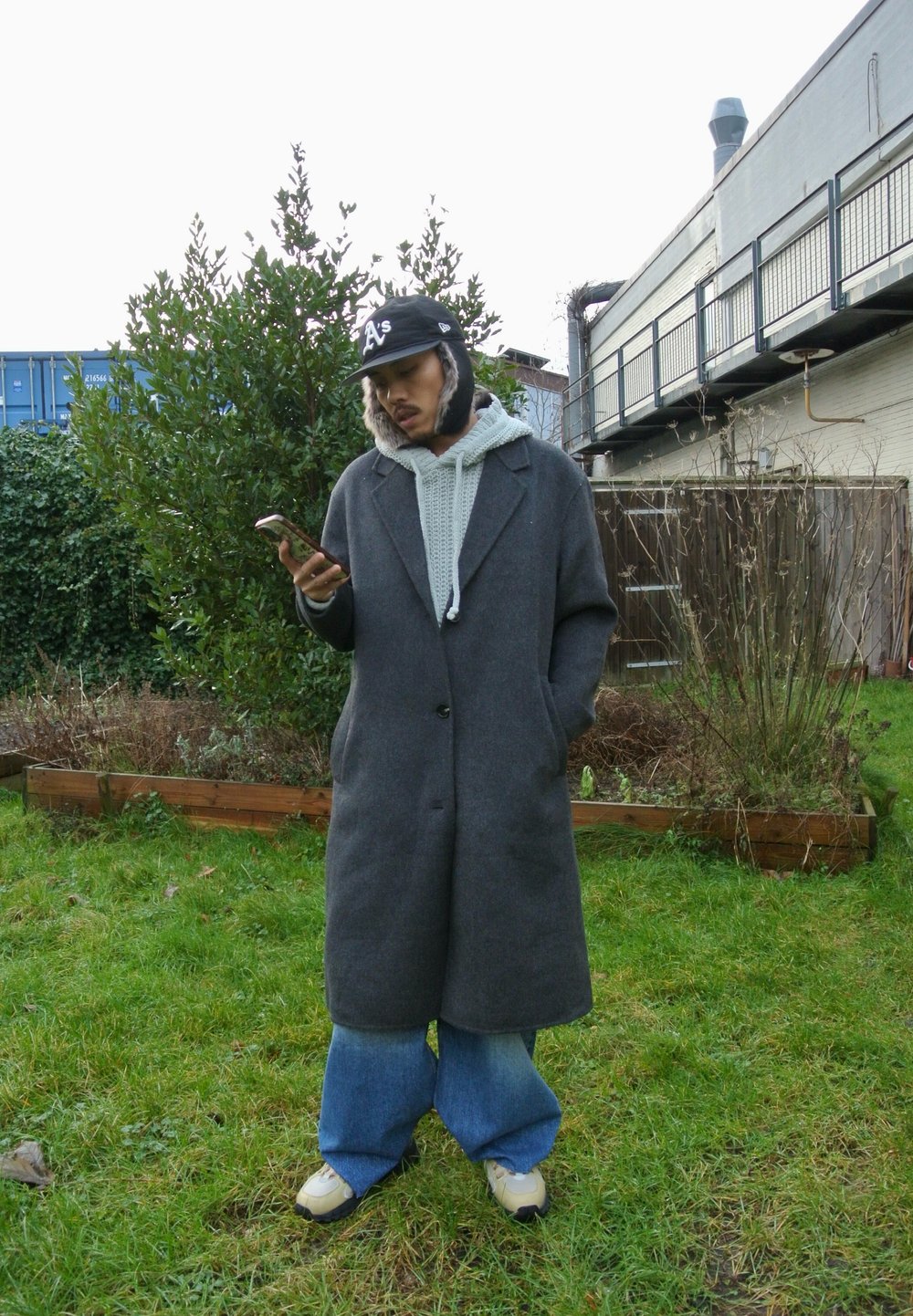 Grigia cappotto di lana con una vestibilità rilassata, abbinato a un hoodie grigio chiaro, jeans blu larghi e sneakers beige. In piedi all'aperto con un telefono.