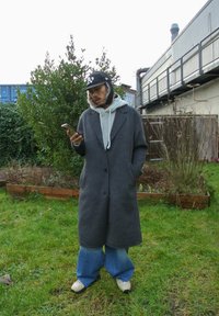 Grigia cappotto di lana con una vestibilità rilassata, abbinato a un hoodie grigio chiaro, jeans blu larghi e sneakers beige. In piedi all'aperto con un telefono.