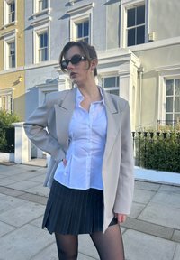Veste oversize beige clair, chemise blanche boutonnée, mini-jupe noire à fines rayures, collants noirs et lunettes de soleil rondes. Fond urbain.