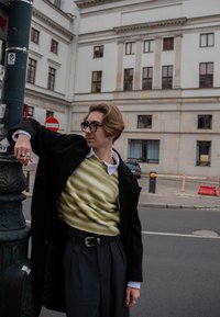 Personne portant un manteau noir par-dessus un pull rayé jaune et crème, un pantalon gris plissé et de grandes lunettes noires, appuyée contre un poteau de signalisation.