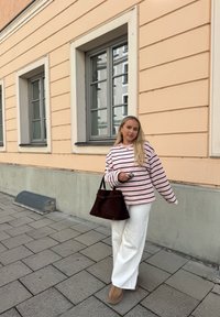 Stribet lyserød og hvid oversize sweatshirt, hvide bukser med vide ben, brun ruskindstaske og brune plateau-støvler foran en pastelvæg.