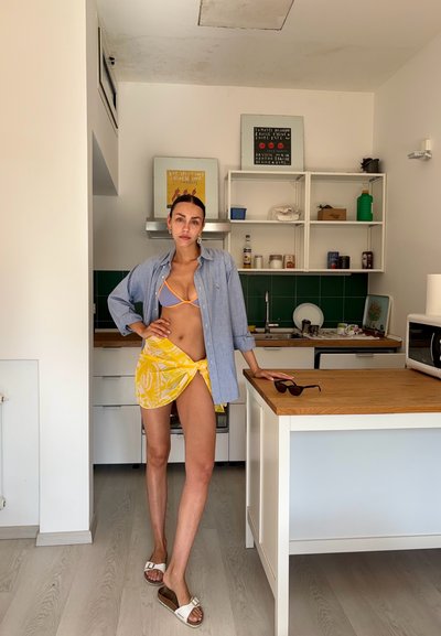 Camisa de botones azul claro, falda envolvente amarilla con estampado, parte de arriba de bikini gris y sandalias blancas; de pie en una cocina moderna con acentos de madera.