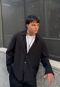 Schwarzes, lässig geschnittenes Hemd mit Knöpfen, über einem weißen T-Shirt getragen, kombiniert mit dunklen Hosen, vor einem strukturierten grauen Hintergrund.