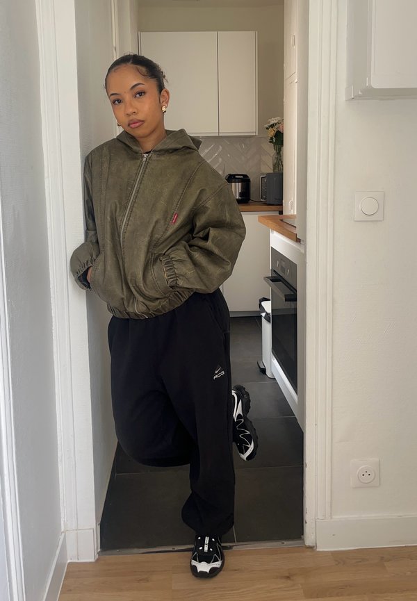 Junge Frau lehnt in einer Küchentür, trägt eine olivgrüne Jacke, schwarze Hose und schwarz-weiße Sneakers.