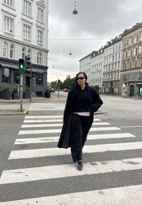 Fekete hosszú kabát fekete kapucnis felső és rózsaszín póló felett, fekete nadrággal és sötét cipővel, egy zebra előtt állva. Az utcán és épületek láthatók.